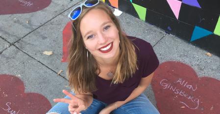 Programomkostninger & hvad er inkluderet Au pair sidder på fortovet, smiler ind i kameraet og laver et fredstegn med hånden.